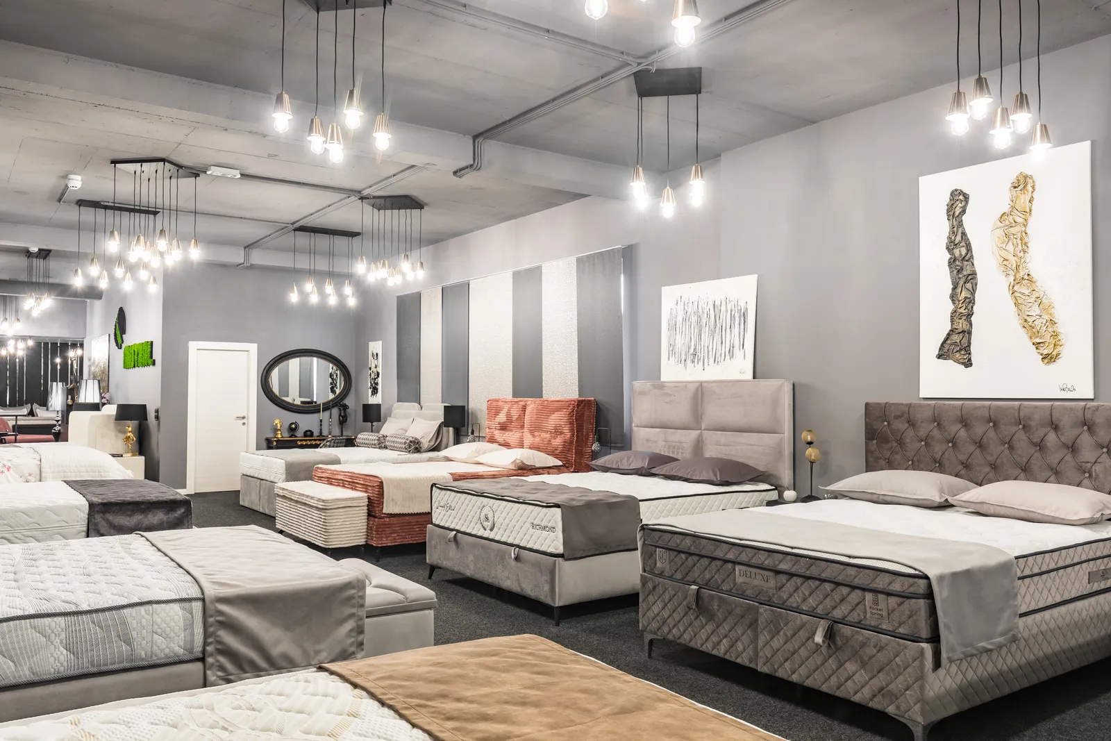 Red izloženih madraca i boxspring ležajeva u salonu, sa slikama i ogledalom na zidu iza.